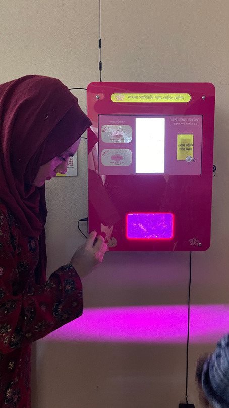 A woman collecting a sanitary pad from a wall-mounted Shapla vending machine